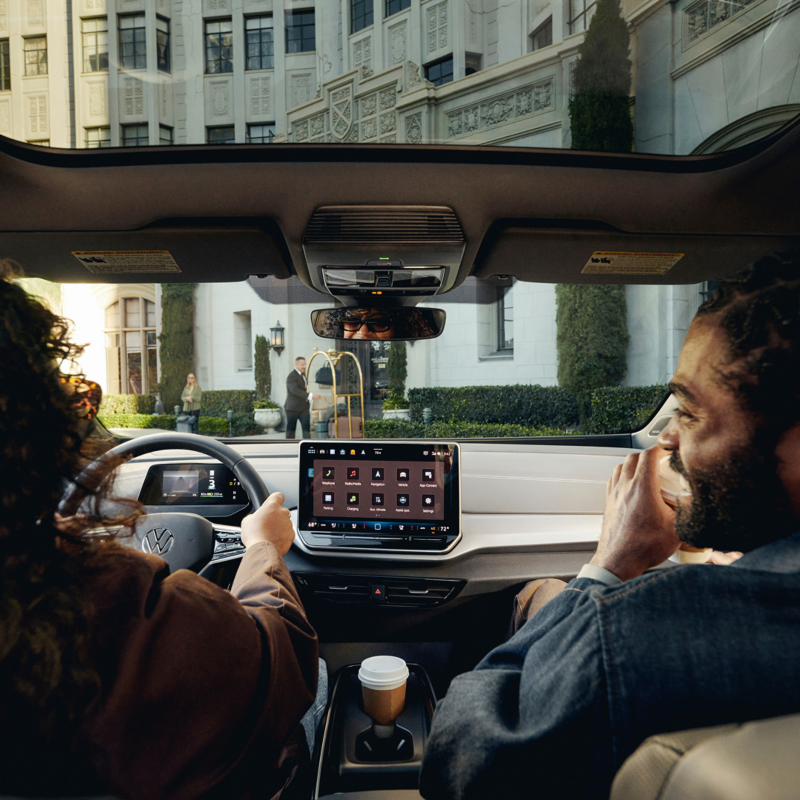 View from inside an ID.4 EV looking out the windshield while a woman drives and a man is smiling in the passenger seat as they approach an upscale hotel. 