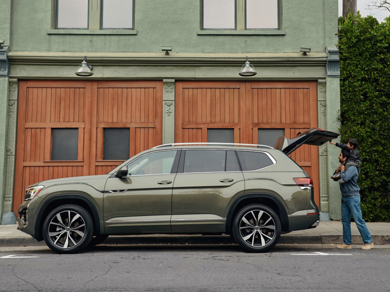 An Atlas shown in Avocado Green Metallic is parked in front of a green building while a dad holds up a child to touch the open liftgate.
