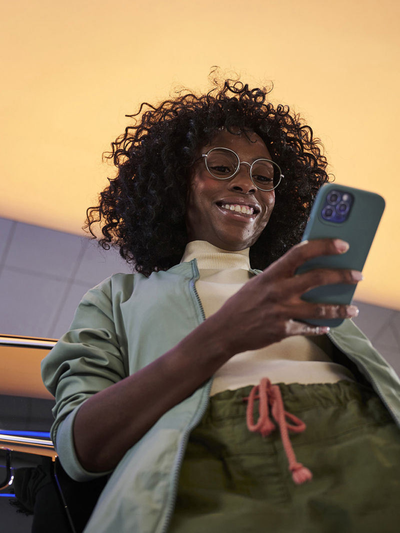 A fashionably dressed woman holds a smartphone while leaning against a car inside a modern lit room.