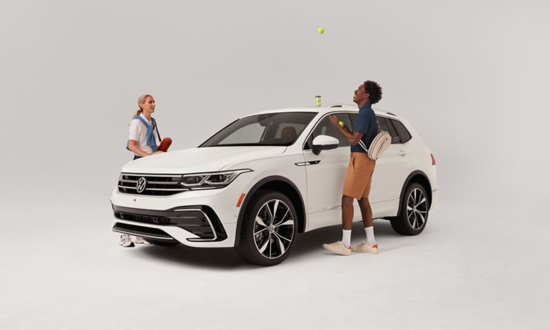 Front ¾ view of a Tiguan shown in Opal White parked on a white background with two people standing outside the vehicle playing with tennis gear. 