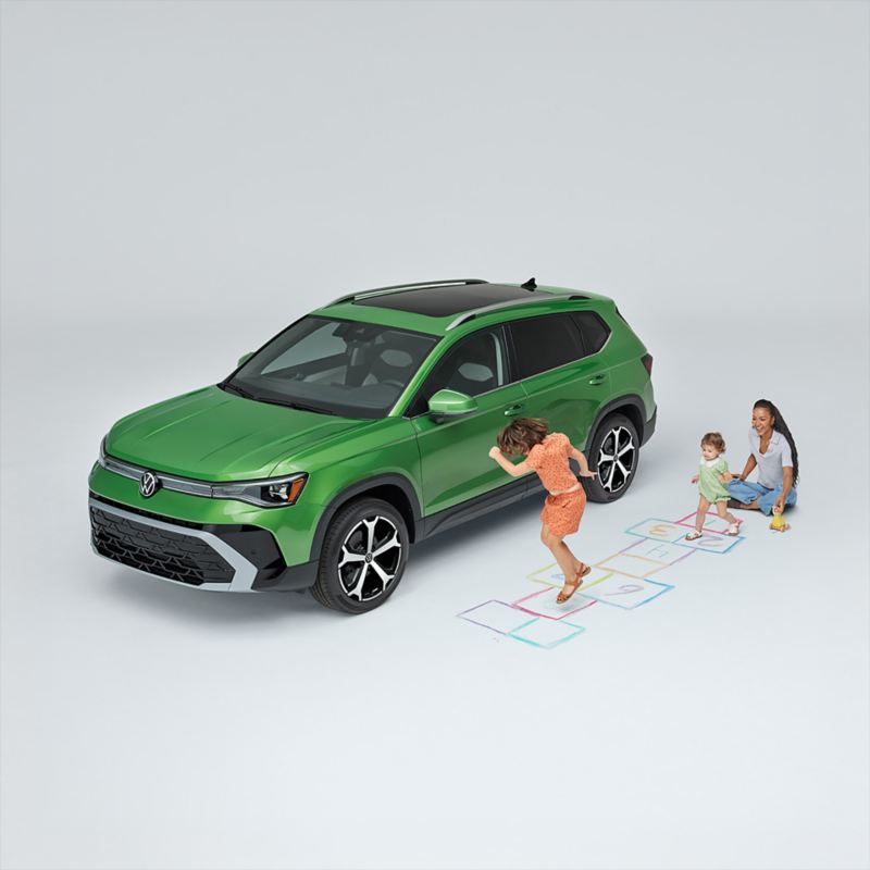 Overhead view of a mom sitting on the ground while children play next to a Taos shown in Bright Moss Green parked on a white sound stage environment.