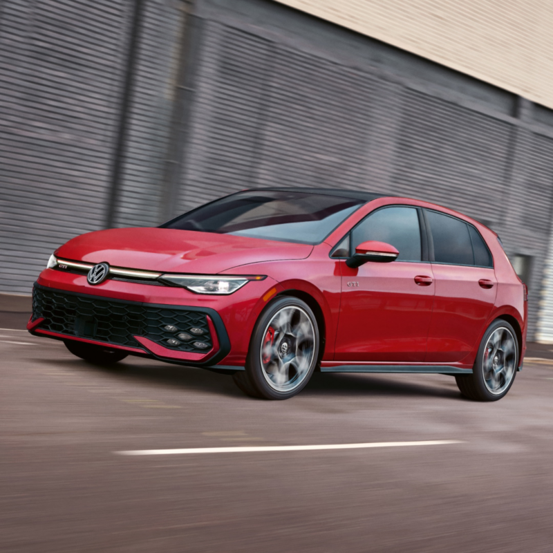 Front view of a Golf GTI shown in Kings Red Metallic driving in an industrial setting.