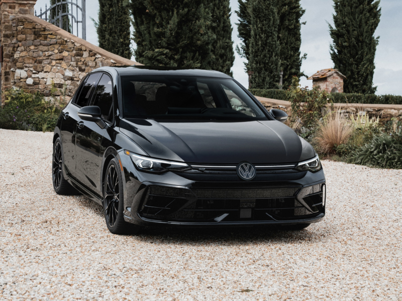 Front view of a Golf R shown in Mythos Black Metallic parked with a low stone wall and bushes in the background.