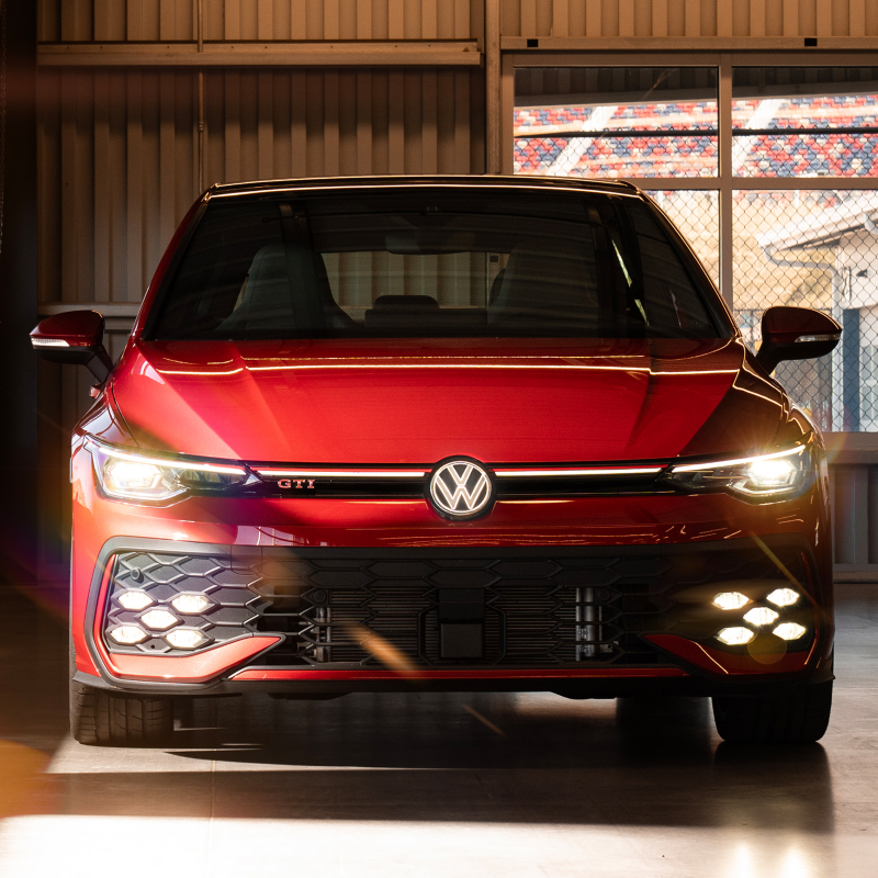 Front view of a Kings Red Metallic Volkswagen Golf GTI parked in a sunlit parking lot.