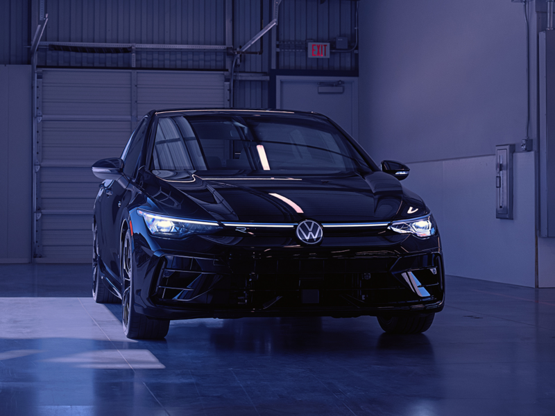 Volkswagen Golf R in Mythos Black Metallic parked in an industrial setting, captured in frontal view with strong reflections and blue-tinted lighting highlighting the vehicle's contours.
