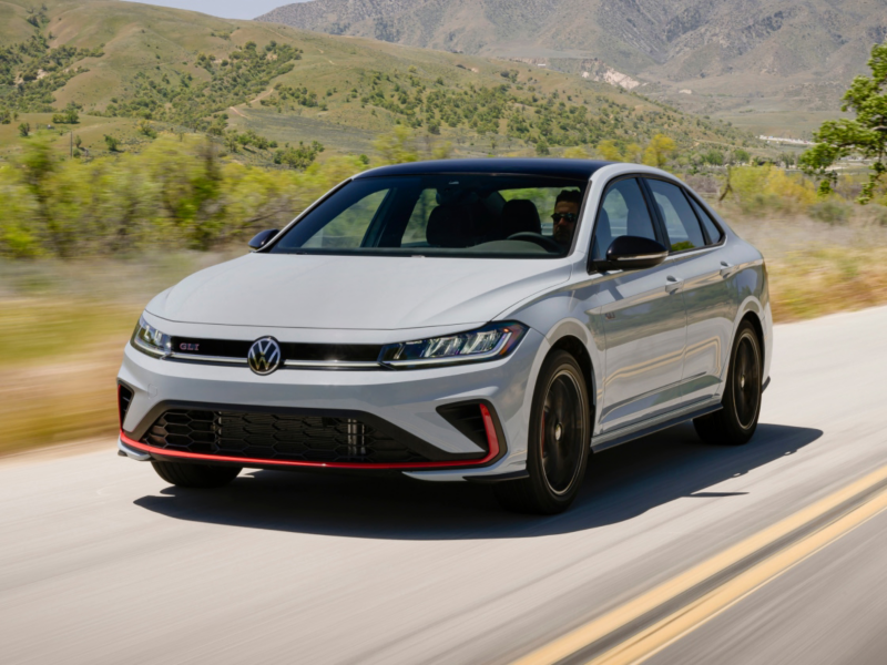 Front 3/4 view of a Jetta GLI shown in Monument Gray driving on a road with a green hill with trees in the background.