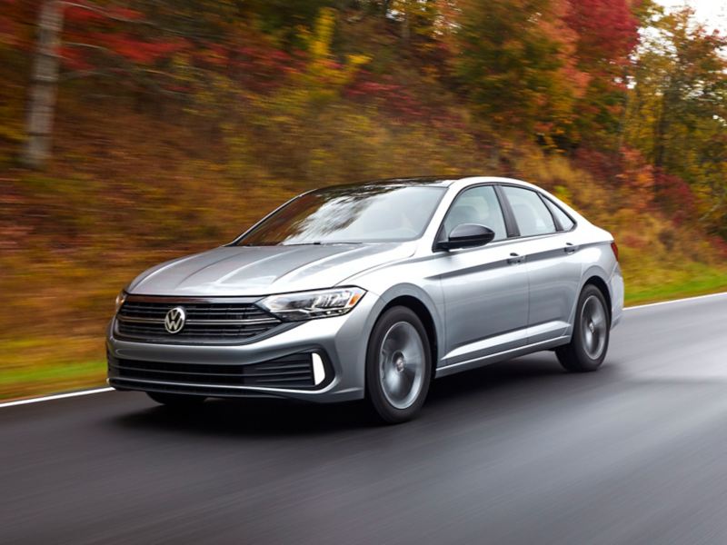 Three-quarter front view of a Jetta shown in Pyrite Silver Metallic, accelerating along a wooded road. Trees show fall colors.