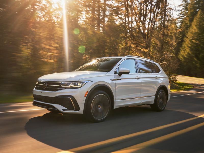 A Tiguan shown in Opal White, driving on a two-lane road with trees in background, as seen from the driver side. 
