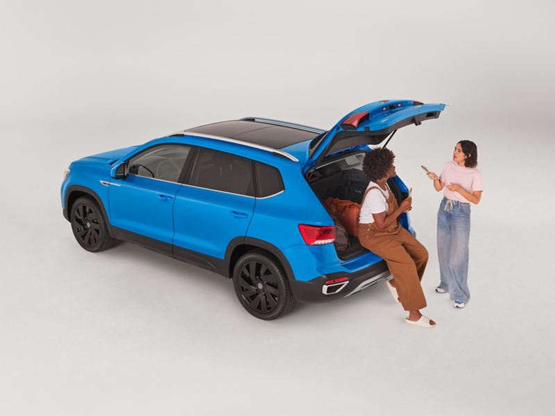 Two people are hanging out and talking at the back of a Taos shown in Cornflower Blue parked on a white background with the rear hatch open with bags in the cargo area.