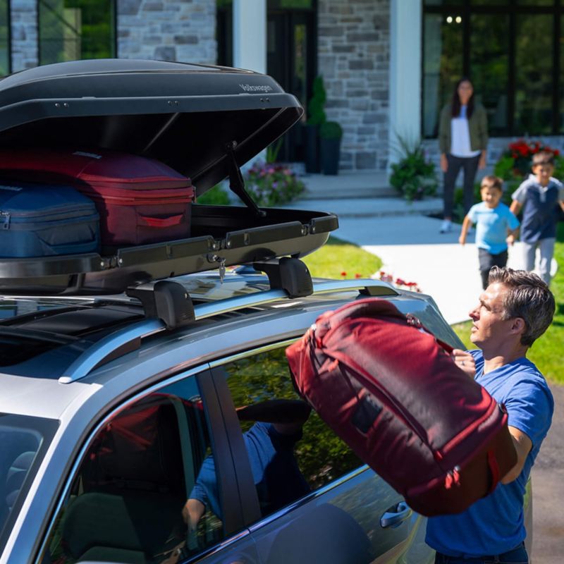 A man loading a duffel bag into a cargo box carrier on a Volkswagen with family and house in the background.