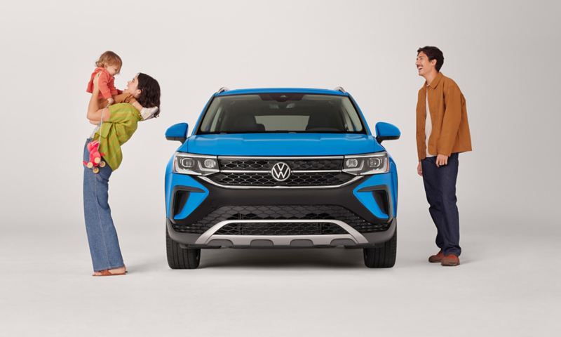 A Taos shown in Cornflower Blue parked on a white studio backdrop with a woman holding a child and a man standing next to the vehicle.