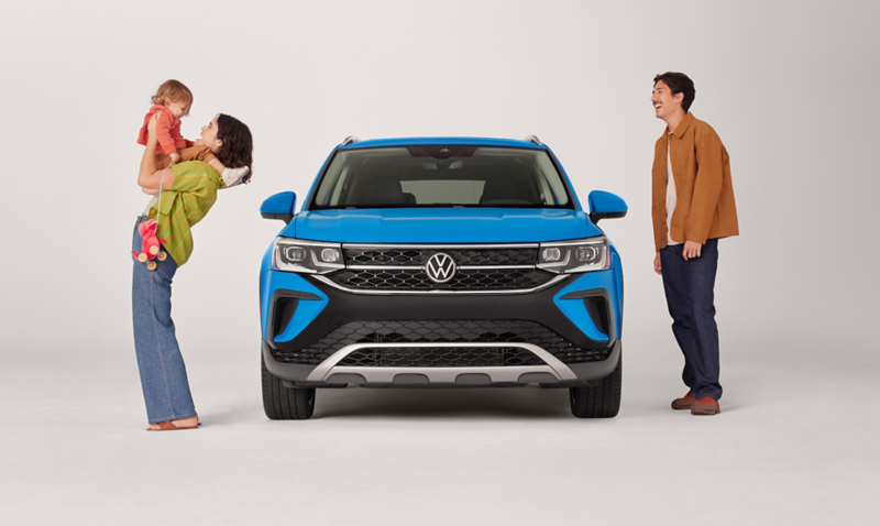 A Taos shown in Cornflower Blue parked on a white studio backdrop with a woman holding a child and a man standing next to the vehicle.