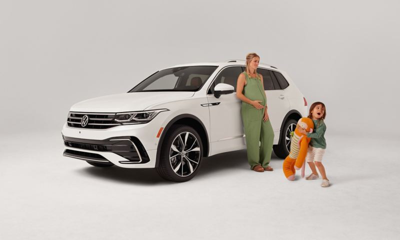 Front ¾ view of a Tiguan in Opal White parked on a white background with a mom leaning on the driver door and a kid holding a stuffed animal.