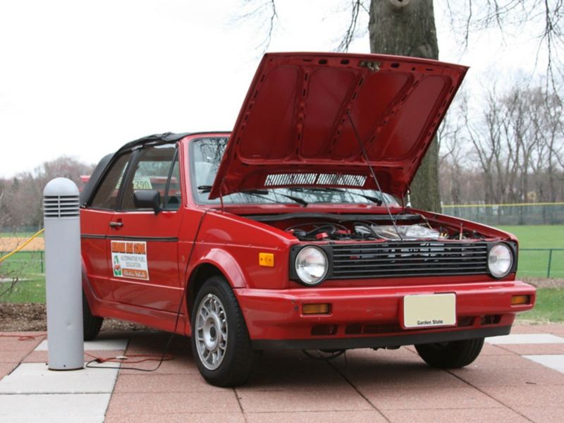 Un Cabriolet Volkswagen rojo 1990 que se ha convertido de gasolina a electricidad está estacionado con su capucha abierta.