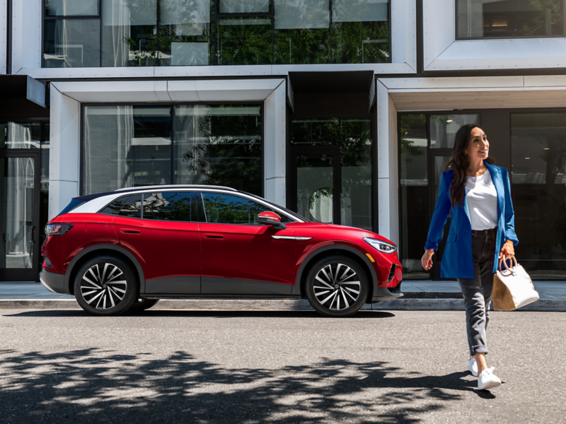 A woman walks away from an Aurora Red Metallic ID.4 SUV that’s parked on an urban street in front of a modern office building.