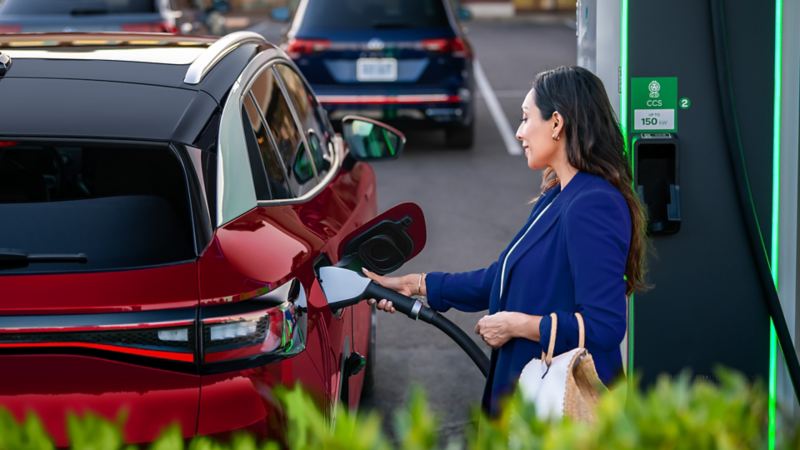 A well-dressed woman, parked at charging station stands on the passenger side of an ID.4 SUV in Aurora Red Metallic plugging a charging cable into the vehicle.