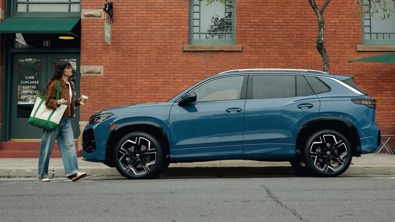 Una mujer que sale de una panadería con una taza de café camina hacia un Tiguan estacionado en color Monterey Blue Pearl con un edificio de ladrillos al fondo.