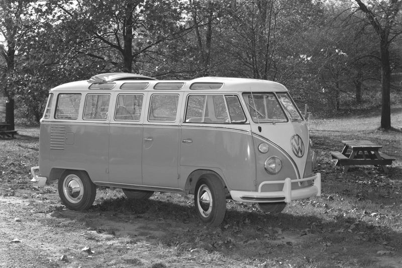 A black and white image of a vintage VW Bus in a park with trees and a picnic table in the background.
