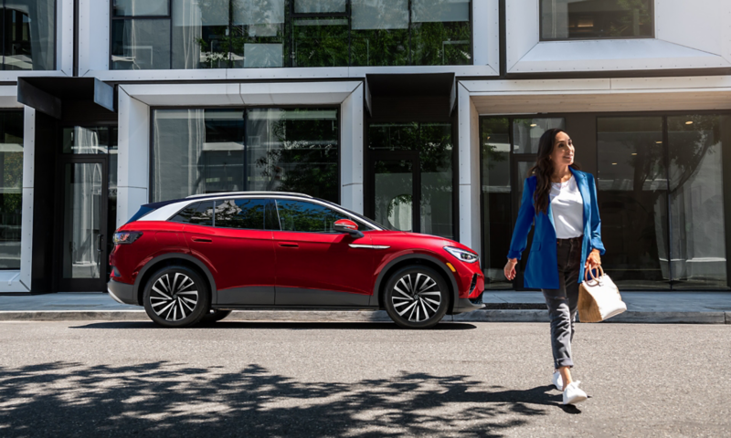 A woman walks away from an Aurora Red Metallic ID.4 SUV that’s parked on an urban street in front of a modern office building.