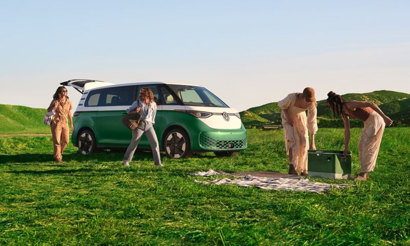 A front ¾ view of the ID. Buzz in Mahi Green Metallic with the rear hatch open and a group of people setting up for a picnic on a grassy field with green rolling hills in the background.