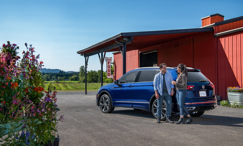 A couple talks at the rear driver side of a Tiguan shown in Atlantic Blue Metallic outside a roadside farm.