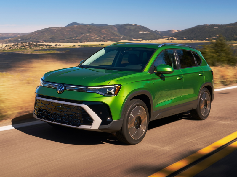 Front 3/4 view of a Taos shown in Bright Moss Green driving with a small lake and mountains in the background.