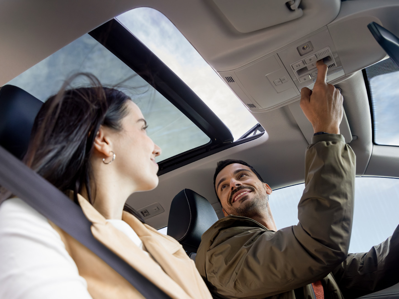 Two smiling people sitting in the front seats of a parked Taos while the driver uses the overhead console controls to open the available panoramic sunroof