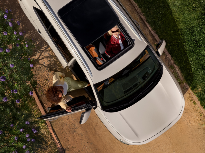 Overhead view of a Tiguan shown in Sandstone parked next to a flowering tree while a women gets into the front passenger seat and the driver is looking up happily through the open available panoramic sunroof