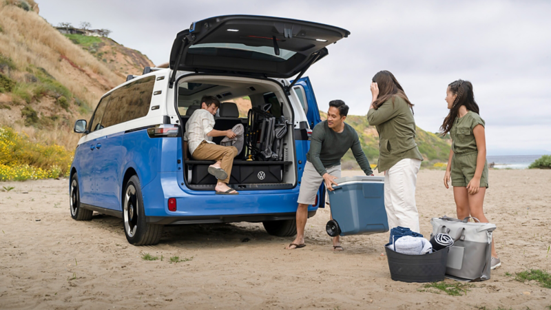 Rear view of an ID. Buzz shown in Cabana Blue Metallic and Candy White parked with a family packing up beach gear using the open rear hatch open and with a beach and ocean in the background.