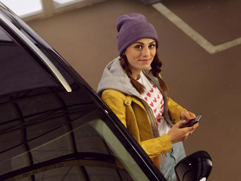 A woman looking at the camera while leaning against a VW holding a phone.