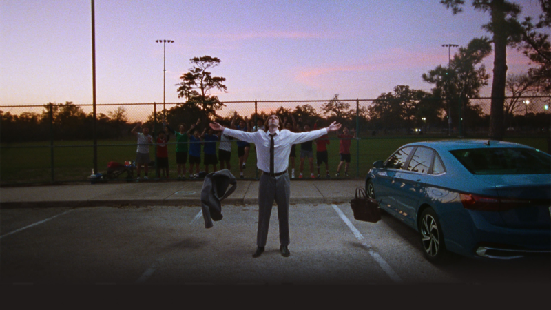 A man standing in a parking lot next to a VW Jetta with a children's soccer team watching him behind a fence in the background.