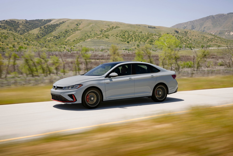 Side angle of a Jetta GLI shown in Monument Gray driving on a two-lane road with wooded hills in the background. 