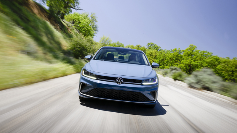 Head-on view of a Jetta shown in Monterey Blue Pearl driving on an open road with trees in the background.