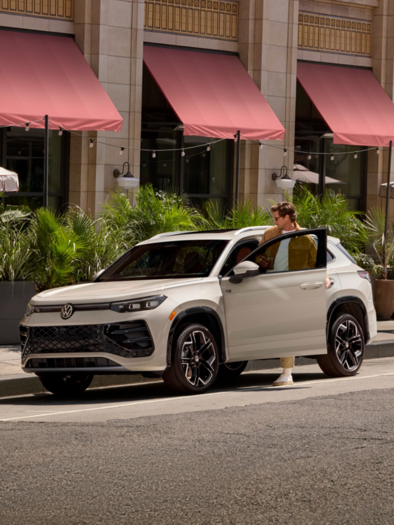 Front 3/4 view of a Tiguan shown in Sandstone parked on an upscale street while a fashionably dressed man enters the vehicle and gets into the driver's seat.