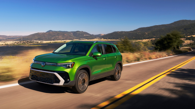 Front view of a Taos shown in Bright Moss Green driving with a small lake and mountains in the background.