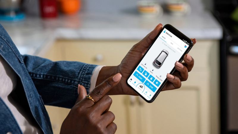 A woman stands in her kitchen holding her phone. The tight crop on the screen, reveals the Car-Net Remote Access page. 