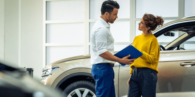 Un técnico de servicio de Volkswagen sosteniendo un portapapeles azul habla con un cliente en un concesionario de VW.