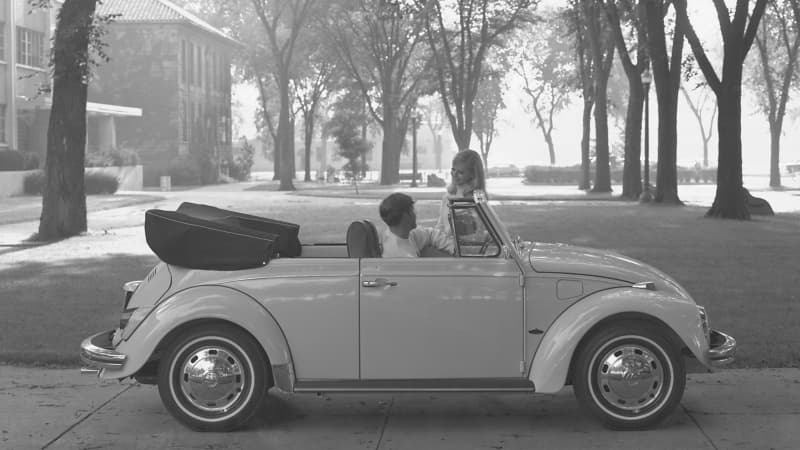 A young man driving a 1969 Volkswagen Beetle convertible, stopped talking to a young woman on a college campus.