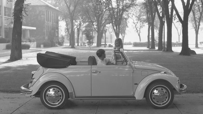 A young man driving a 1969 Volkswagen Beetle convertible, stopped talking to a young woman on a college campus.