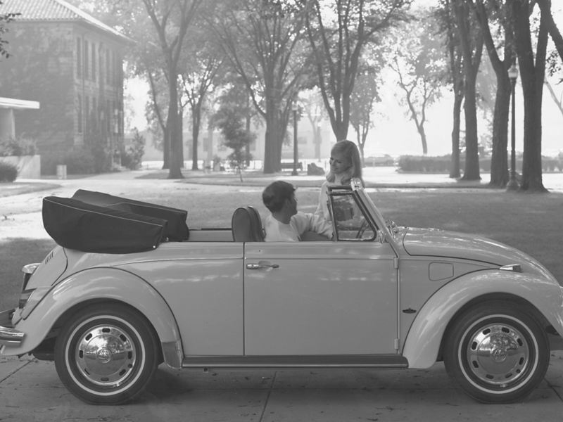A young man driving a 1969 Volkswagen Beetle convertible, stopped talking to a young woman on a college campus.