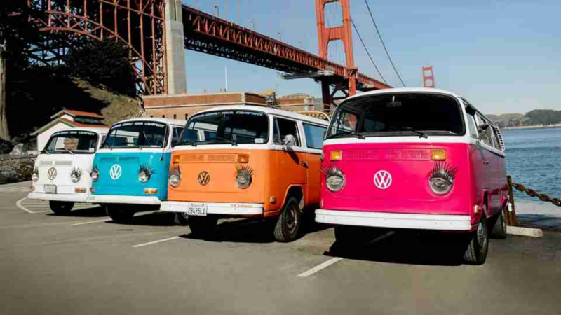 Four vintage Volkswagen buses sit in front of a bridge