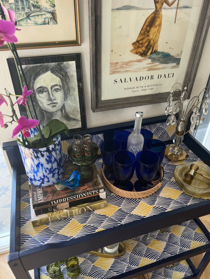 The top of a bar cart, which holds books, a bottle, glassware, and other items.