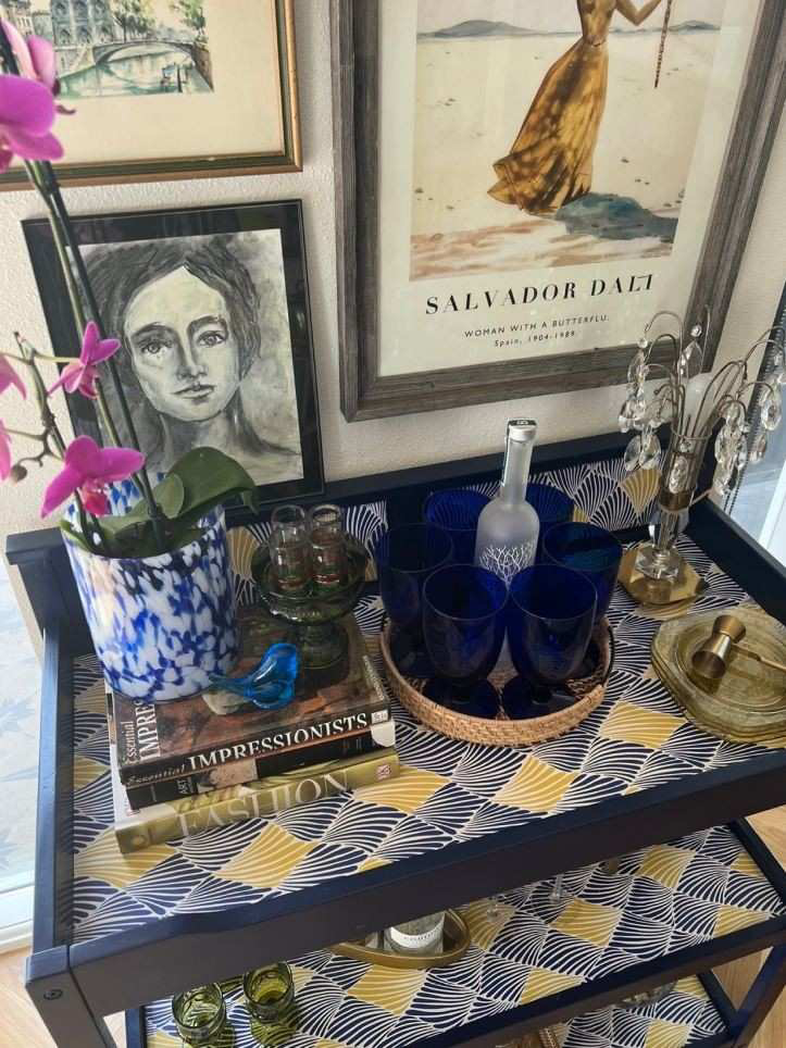The top of a bar cart, which holds books, a bottle, glassware, and other items.