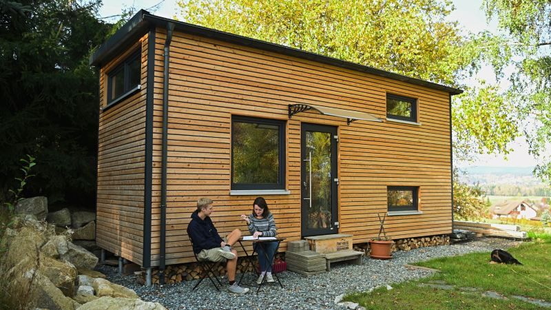 Two people sit at a table outside a tiny house.