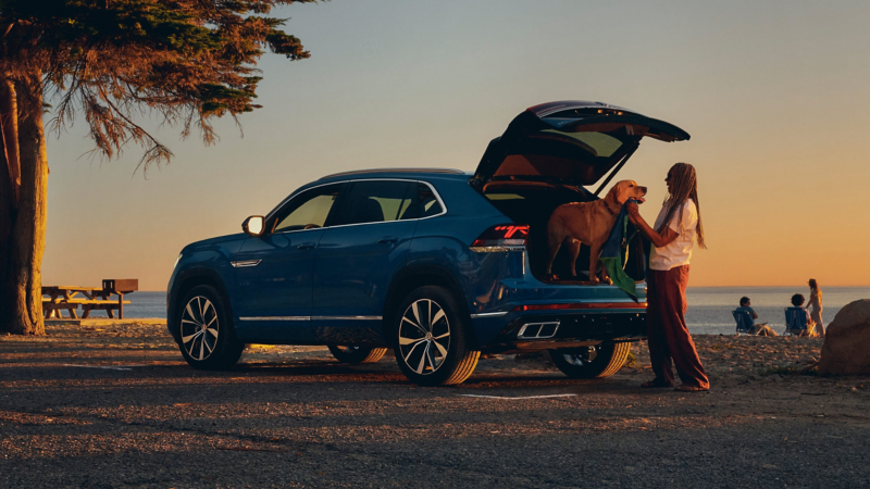 Un Atlas Cross Sport mostrado en Mountain Lake Blue Metallic está parqueado en una playa al atardecer mientras una mujer emplea la zona de carga abierta para secar con toalla a su perro grande.