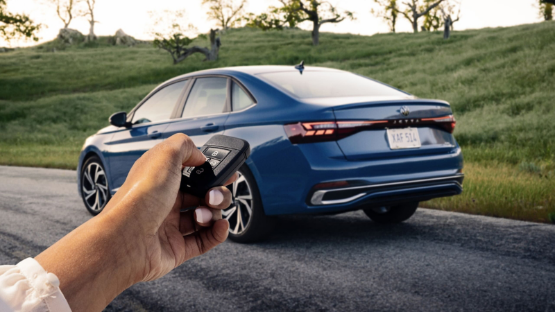 Rear view of a Jetta shown in Monterey Blue Pearl parked with trees in the background and a person holding a VW key fob in the foreground