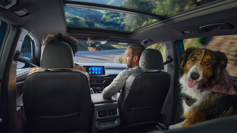 Interior view of a dog in the back row looking at the camera and the front seats with passengers of an Atlas Cross Sport driving on a road with mountains in the background.