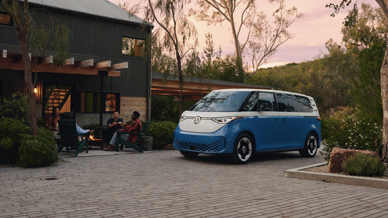 An ID. Buzz shown in Cabana Blue Metallic and Candy White is parked in front of a modern house while a group of friends gather around a fire pit eating snacks on the house’s patio.