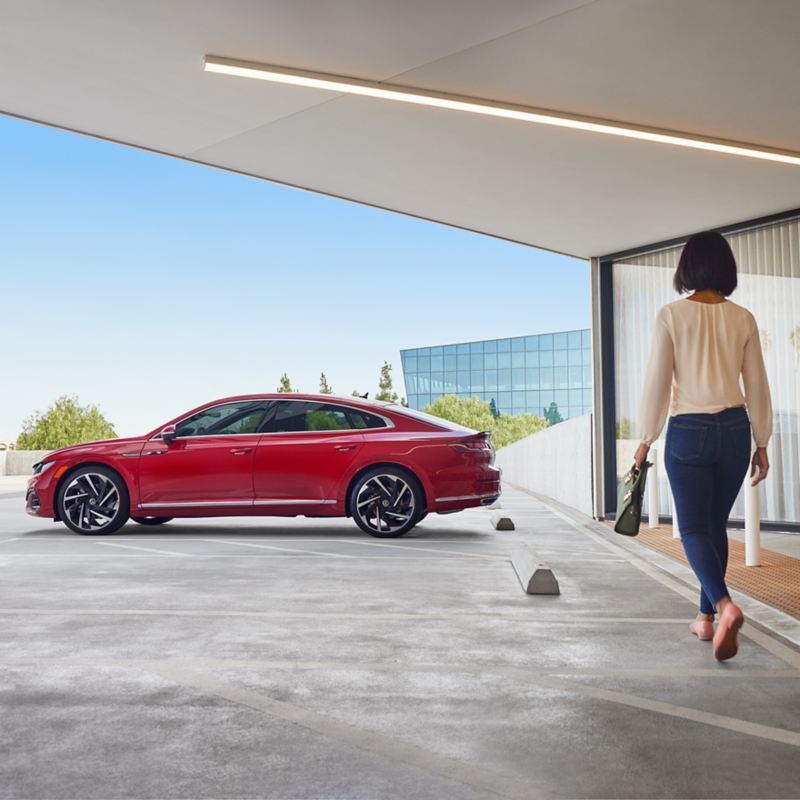 A woman approaches an Arteon shown in Kings Red Metallic parked on the upper-level of a parking garage in a modern building.