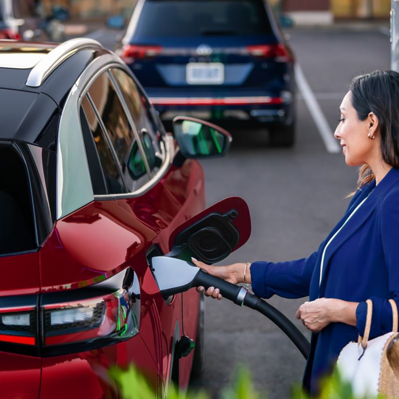 A well-dressed woman, parked at charging station stands on the passenger side of an ID.4 SUV in Aurora Red Metallic plugging a charging cable into the vehicle.